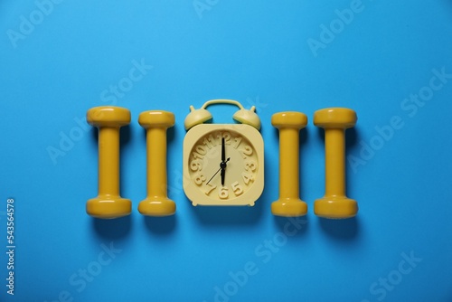 Yellow alarm clock and dumbbells on light blue background, flat lay. Morning exercise