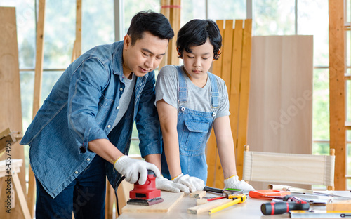 Asian cheerful male dad son carpenter woodworker colleague in jeans outfit with safety gloves helping using pencil drawing writing blueprint on working workshop table in housing construction site