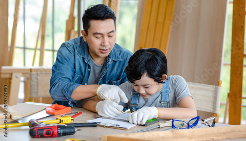 Asian cheerful male dad son carpenter woodworker colleague in jeans outfit with safety gloves helping using pencil drawing writing blueprint on working workshop table in housing construction site