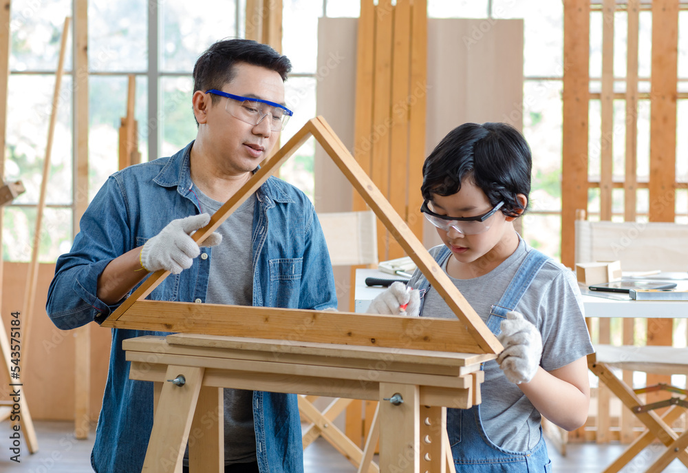 Asian male carpenter woodworker engineer dad in jeans outfit with safety gloves and glasses goggles helping teaching son using pencil marking wood stick board plank on workbench in construction site Asian male carpenter woodworker engineer dad in jeans outfit with safety gloves and glasses goggles helping teaching son using pencil marking wood stick board plank on workbench in construction site