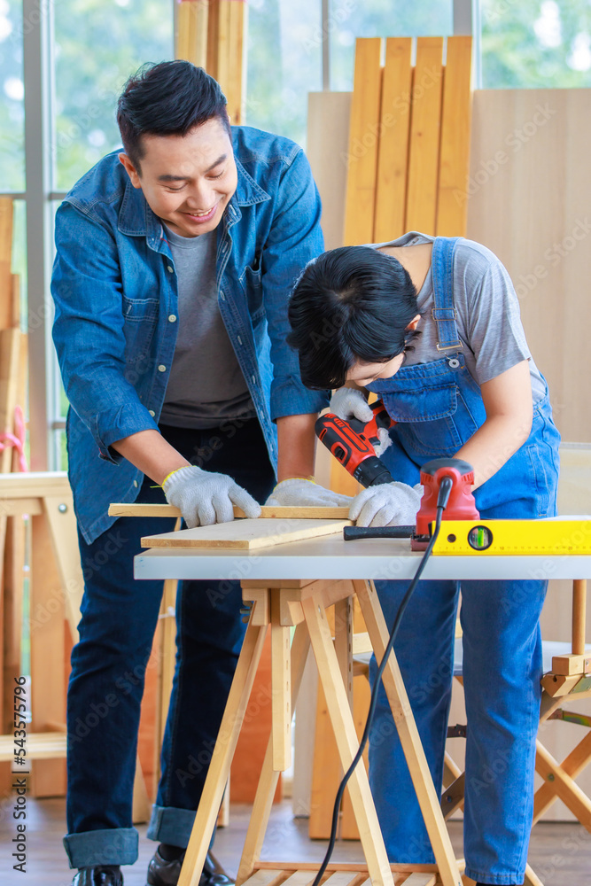 Asian cheerful male dad and son carpenter woodworker colleague in jeans outfit smiling helping using drill machine digging hole in wood stick on working workshop table in housing construction site