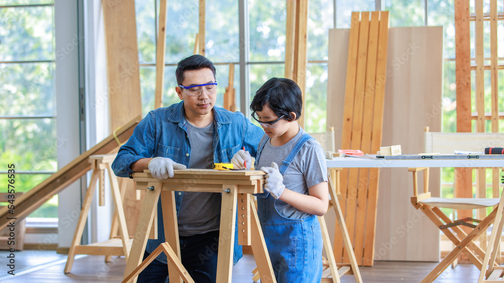 Asian male carpenter woodworker engineer dad in jeans outfit with safety gloves and glasses goggles helping teaching son using pencil marking wood stick board plank on workbench in construction site