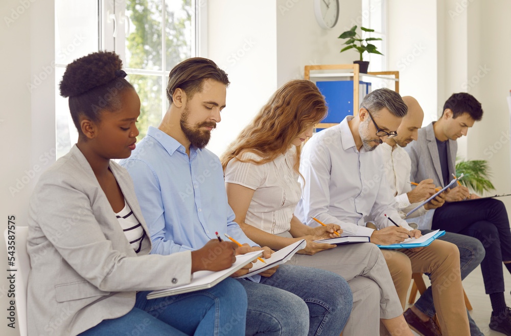 Group of international business people different ages studying, writing making notes listening presentation in office sitting on chairs during meeting or business training. Adult education concept. Group of international business people different ages studying, writing making notes listening presentation in office sitting on chairs during meeting or business training. Adult education concept.