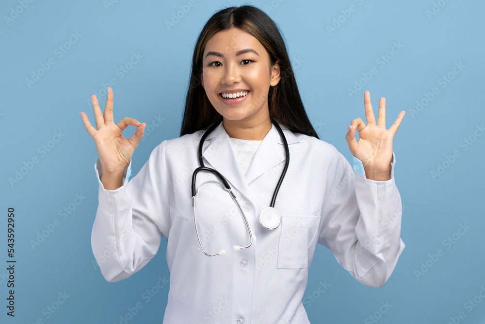 Portrait of satisfied asian woman doctor standing, looking at camera showing Ok sign