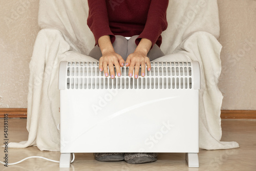 A girl sits on an armchair and puts her hands on a warm convector in an apartment in winter, a heating season, a warm house