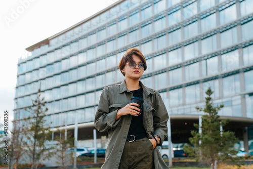 Wallpaper Mural Indifferent young Asian woman with short hair holding coffee cup and hand in pants pocket standing against glass building and looking away Torontodigital.ca