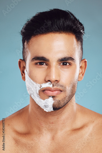 Portrait, skincare and man shaving in studio on blue background for facial hair grooming in India. Healthy, products and young face model shaves beard and mustache in self care routine with cream