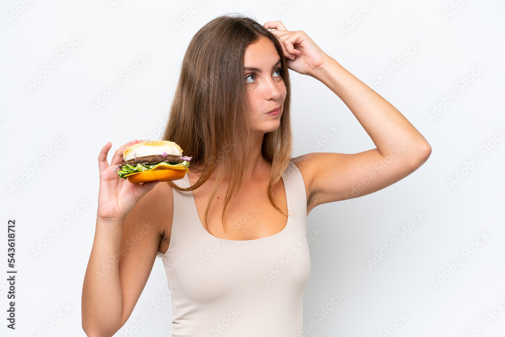 Young pretty woman holding a burger isolated on white background having doubts and with confuse face expression Young pretty woman holding a burger isolated on white background having doubts and with confuse face expression