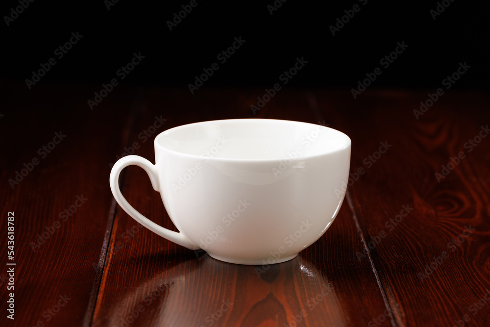 A white, clean, busty china cup against a dark wooden table. Side view. A white, clean, busty china cup against a dark wooden table. Side view.