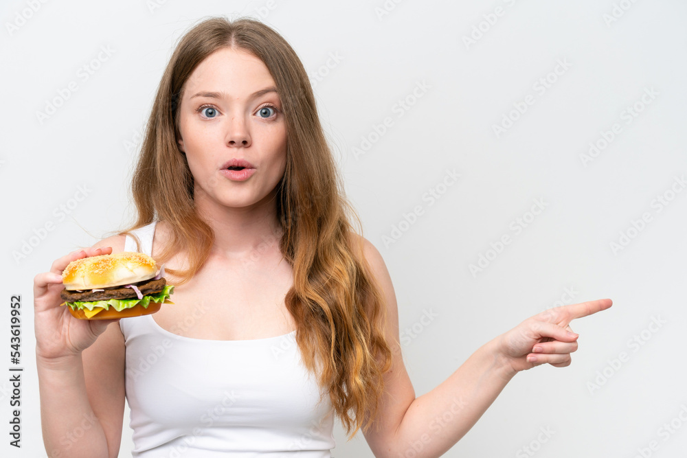 Young pretty woman holding a burger isolated on white background surprised and pointing side Young pretty woman holding a burger isolated on white background surprised and pointing side