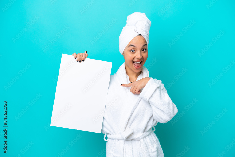 Young woman in a bathrobe with towel isolated on blue background holding an empty placard with happy expression and pointing it Young woman in a bathrobe with towel isolated on blue background holding an empty placard with happy expression and pointing it