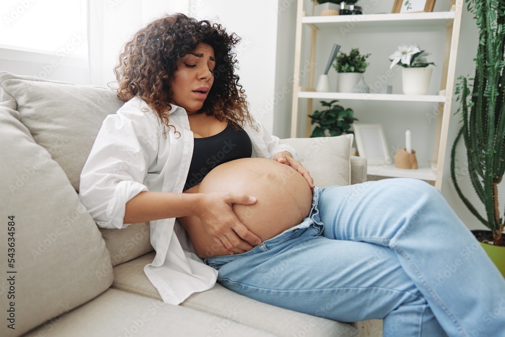 Pregnant woman headache lies at home in a shirt and jeans on the couch fatigue and heaviness in the last month of pregnancy before childbirth, lifestyle difficulties of motherhood, nausea Pregnant woman headache lies at home in a shirt and jeans on the couch fatigue and heaviness in the last month of pregnancy before childbirth, lifestyle difficulties of motherhood, nausea