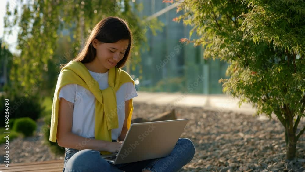 Asian student sitting on the bench studying on the laptop in the city centre. Female student studying, using laptop for studying purposes. People and technology concept