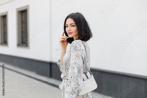 Fashion beautiful woman with brunette hairstyle in fashionable flowers spring dress with stylish white bag walks on the street and looks at the camera