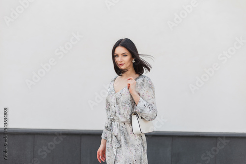 Beautiful young fashion girl in stylish elegance dress with flowers print with trendy fashion handbag stands near a white wall on summer windy day photo