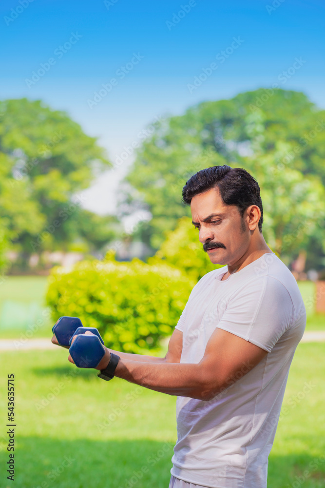 Young indian man doing exercise by lifting dumbbells in the park.