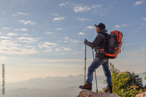 Male hiker holding stick with backpack standing on top mountain rock and enjoying sunset. Hiker men's hiking living healthy active lifestyle