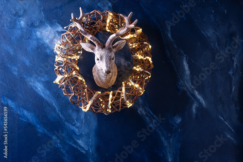Symbol of Christmas and New Year -  handmade  rustic wreath with  bright fairy ligts and head of deer on deep blue  textured wall. Place for text. Christmas postcard. photo