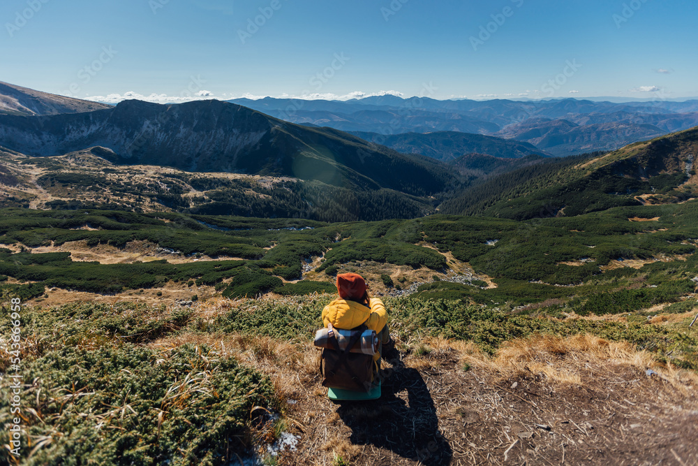 Woman hikerin yellow down jacket with backpack is sitting on stunning panoramic view of mountains in sunny autumn day. Active vacations concept image