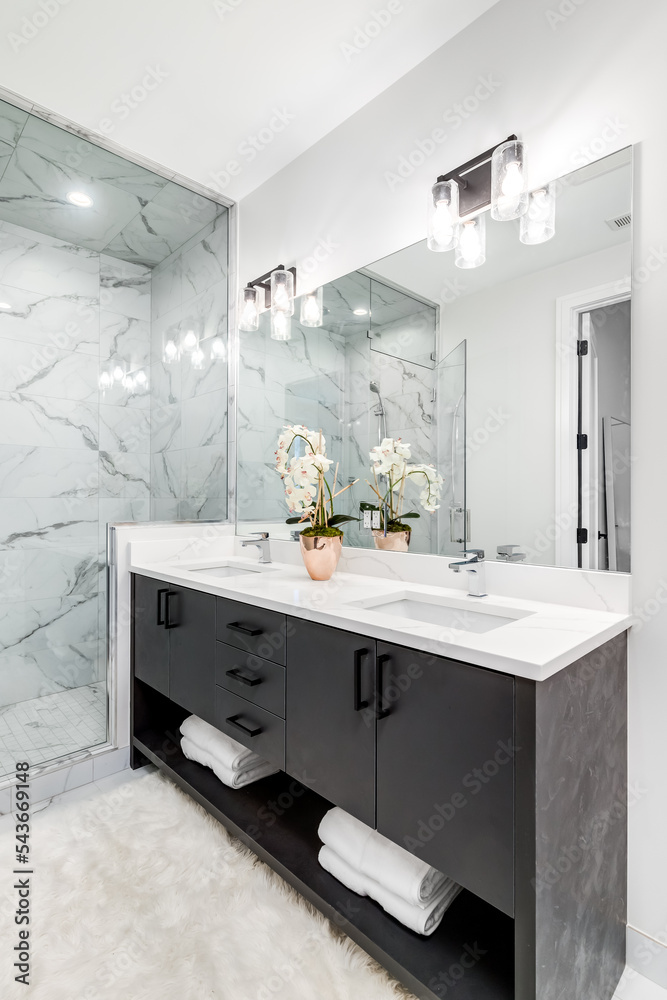 A beautiful bathroom with a dark vanity and white granite counter top. A  plant and towels sit on the vanity. The shower is lined with marble tiles.  Lights on. Photos | Adobe