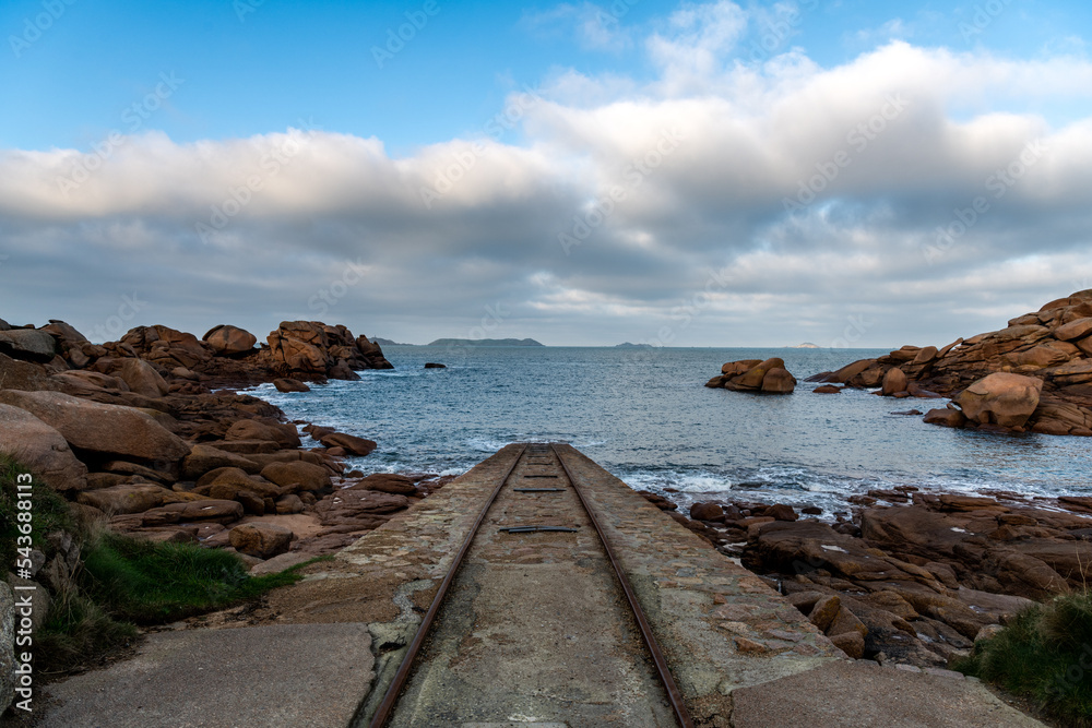 France, Ploumenach, 2022-01-13. Rails for launching boats run into the water along the pink granite coast in Brittany with the tip of the Mean Ruz lighthouse. 