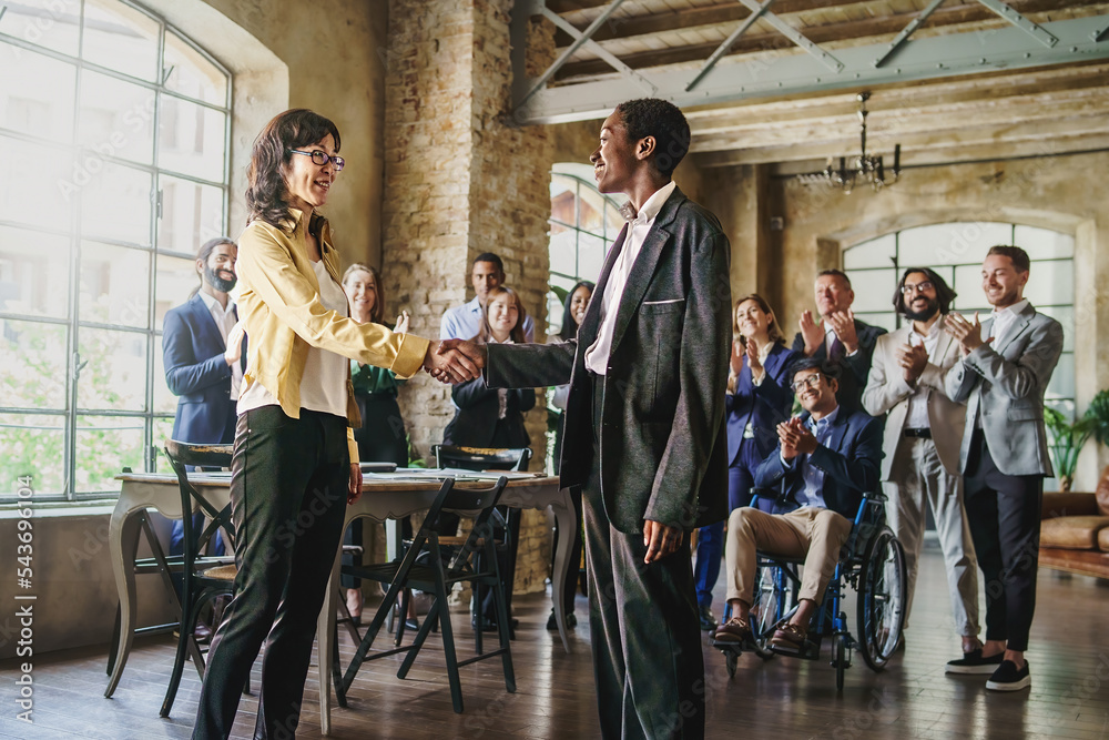 Obraz premium Multiethnic businesswomen shaking hands in front of their colleagues applauding in background - business lifestyle concept