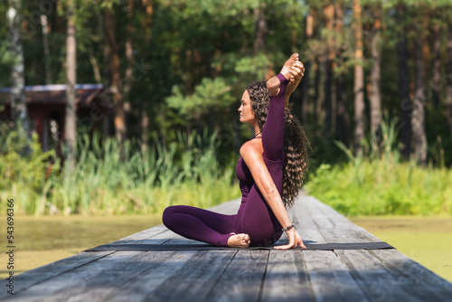 Woman leading a healthy lifestyle and practicing yoga, performs a variation of the yantrasana exercise, compass pose, trains in sportswear in the park, sitting on a wooden bridge photo