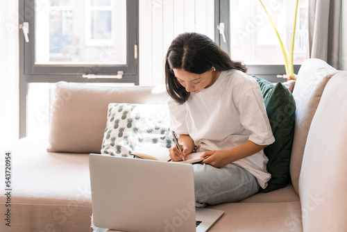 Charming brunette girl making notes while sitting on cozy couch at home during the weekend