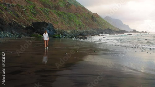 Woman tourist walks along the black volcanic sand beach at sunset. Canary island, Tenerife. Rear view. Relax and enjoy treveler concept. Night nature. photo