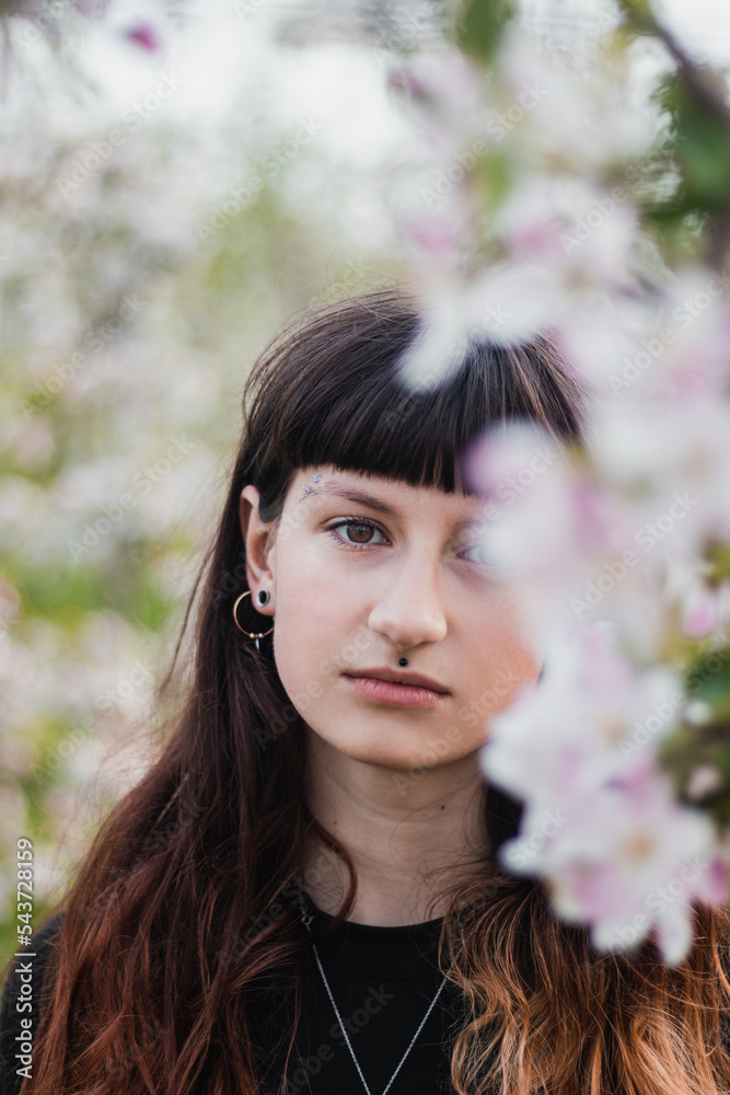 Candid portrait of a beautiful brown-haired woman with facial piercings in a blossoming apple orchard. Brown-eyed girl with cute face Candid portrait of a beautiful brown-haired woman with facial piercings in a blossoming apple orchard. Brown-eyed girl with cute face