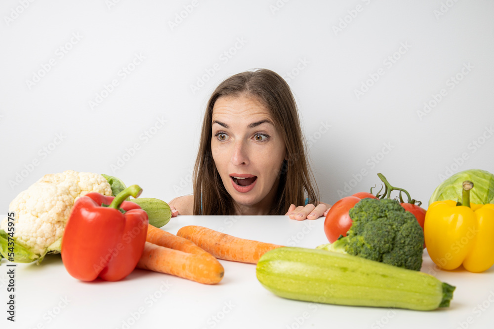 Young woman peeks out from under the table in surprise looking at fresh vegetables Young woman peeks out from under the table in surprise looking at fresh vegetables