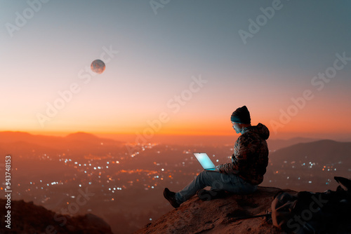 Fototapeta silhouette of digital nomad sitting on top of a hill working with his laptop ove