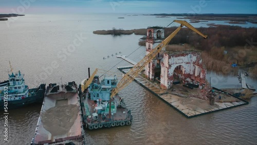 A crane on a floating platform unloads sand to strengthen the shore around the old ruined lighthouse. Aerial drone shot photo