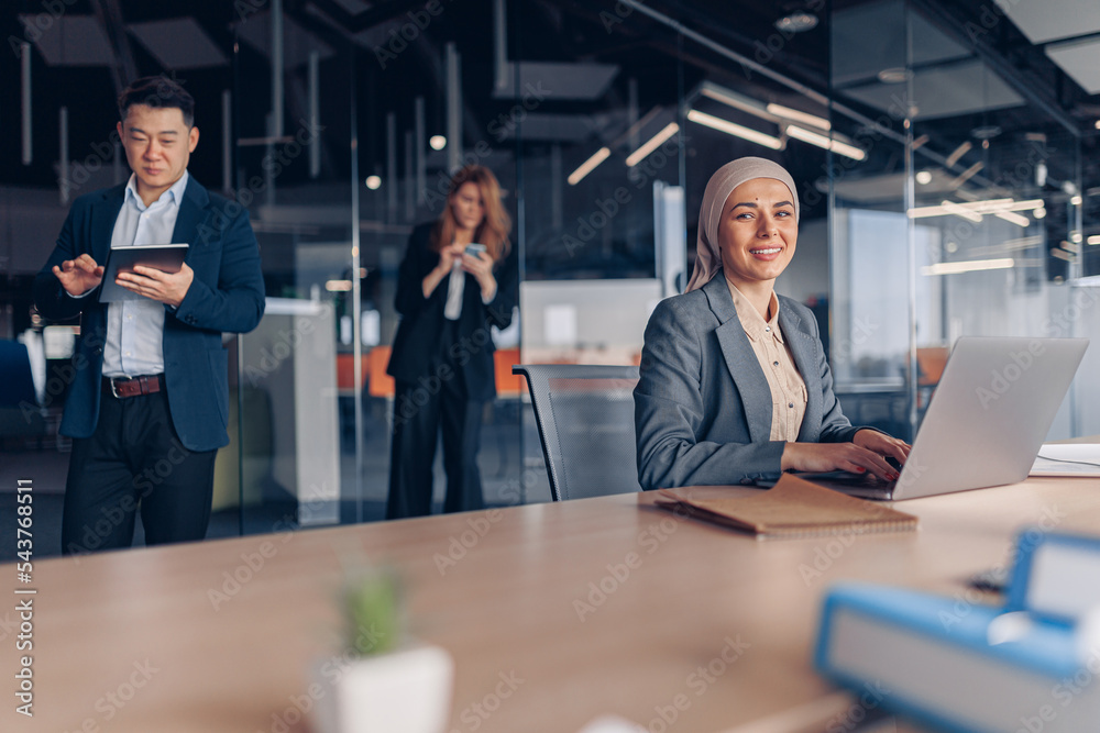 Smiling muslim businesswoman in hijab working on computer while sitting in modern office