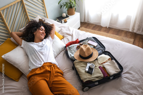Fotografija Happy woman lying down on bed next to open suitcase full of clothes, passport and hat, ready to go on vacation trip