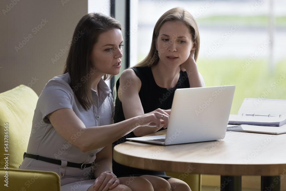 Two focused businesswomen, talking, discuss on-line project, learn collaborative task looking at laptop screen seated in modern office, mentor explaining new corporate software. Workflow, modern tech Two focused businesswomen, talking, discuss on-line project, learn collaborative task looking at laptop screen seated in modern office, mentor explaining new corporate software. Workflow, modern tech