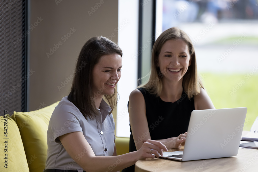Two smiling attractive businesswomen sit at table look at laptop, workmates watch on-line videos, interesting creative content during break at workplace. Workflow using modern technology and internet