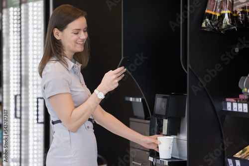 Young attractive businesswoman holding smart phone while prepare morning hot beverage using professional coffee vending machine standing in office workspace during lunch break chatting with friend.