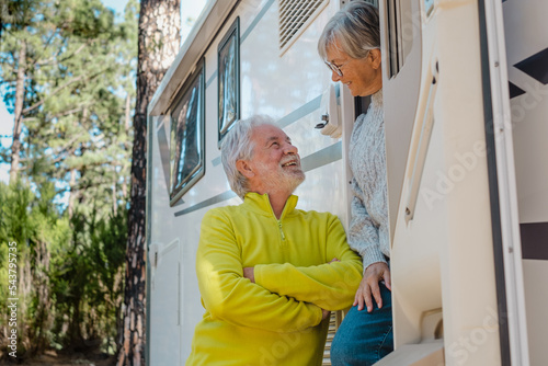 Happy relaxed senior couple on the door of their camper van motor home while talking. Smiling attractive elderly people enjoying retirement in freedom vacation travel in the forest.