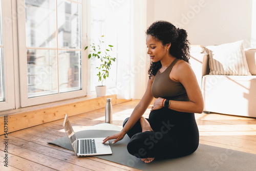 African american pregnant woman looking at laptop screen, searching for online workout tutorial or prenatal yoga sitting on floor on mat in sports wear, wearing smart watch to control heart rate photo
