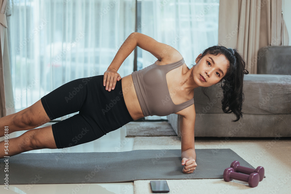 Obraz premium Asian woman exercise at home. Healthy female in sportswear doing side plank workout for abs muscles in living room, Health care and wellness concept.