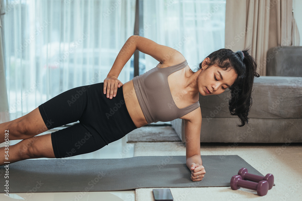 Obraz premium Asian woman exercise at home. Healthy female in sportswear doing side plank workout for abs muscles in living room, Health care and wellness concept.