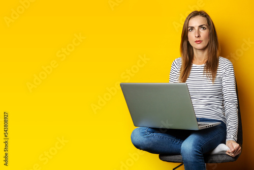 Young woman sitting on chair and holding laptop on isolated yellow background.