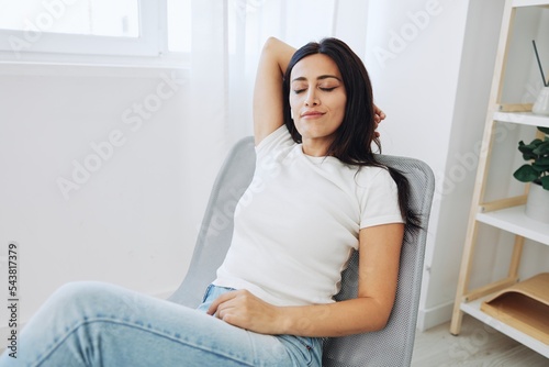 Woman sitting in a chair listening to an audiobook on wireless headphones at home in jeans and a white T-shirt, fall lifestyle comfort