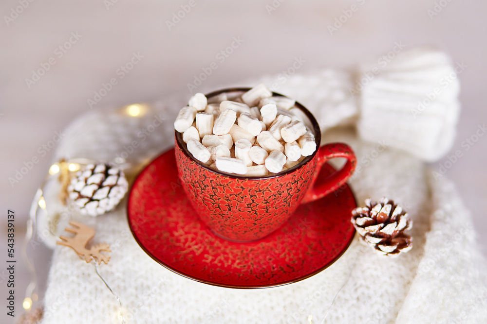 A red cup of cocoa with marshmallows on a white knitted sweater, a garland with Christmas decorations. Christmas winter composition, cozy home atmosphere. A red cup of cocoa with marshmallows on a white knitted sweater, a garland with Christmas decorations. Christmas winter composition, cozy home atmosphere.