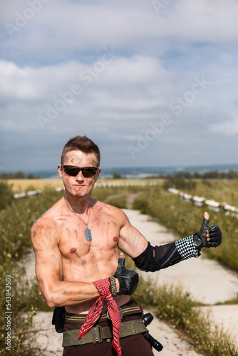 male vandal with dirty naked torso shows like gesture with his hands, against background of an overgrown abandoned road