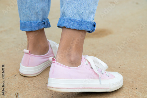 A rear view of a teenage girl's ankle in pink sneakers and blue jeans against a background of yellow dry grass. The concept of clothing for modern teenagers in the cool season