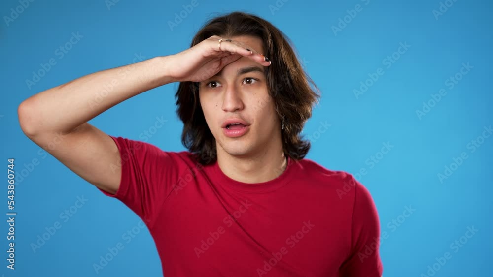 Portrait of happy smiling Hispanic gender fluid young man 20s smiling and look around for friend find waving meet greet with hand as notices someone isolated on blue background.