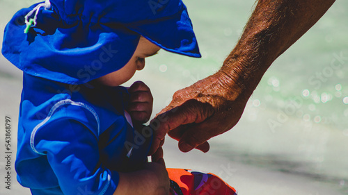 Grandfather's mature hand pointing at belly button of grandson, young toddler at the beach in florida with ocean water in background during summer