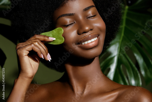 Gua Sha jade skin treatment. African American model making face massage  with a stone tool against tropical green leaves background. photo